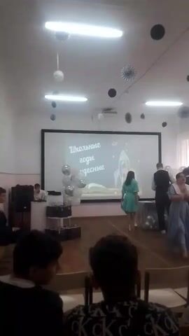 A medium shot captures an indoor event, possibly a school assembly or graduation ceremony, in a room with a large projection screen at the front. The screen displays text in Russian that translates to "School years are wonderful" and features a stylized drawing of a rocket and clouds. In front of the screen, a young woman in a teal dress is standing near a decorated table with several silver balloons. A group of young men in formal attire, including suits and vests, are gathered around the table, some appearing to be interacting with the woman. To the left of the stage, there are stacks of what appear to be black briefcases or boxes. The room is illuminated by fluorescent lights on the ceiling, and decorative black and silver paper fans are hung around the room. In the foreground, the backs of several audience members are visible, seated and facing the stage. The overall atmosphere suggests a celebratory or commemorative event.