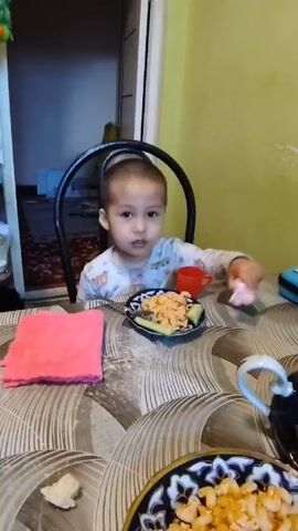 A young child sits at a table ready to eat a meal. The child has short hair and is wearing a patterned pajama top. On the table in front of them is a plate with macaroni, a slice of cucumber, a pink cloth, a small red cup, and a pink toy. There is another bowl of macaroni on the table. The child looks directly at the camera. The table is patterned with various shades of gray. The wall is yellow. A doorway is visible, leading to another room.