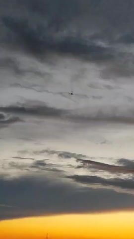 A small propeller plane is silhouetted against a dramatic cloudy sky during sunset. The sun creates a vibrant orange glow along the horizon, illuminating the undersides of the clouds with hues of pink and gold. Darker, heavier clouds occupy the upper portion of the frame, creating a stark contrast with the brighter sky below. The plane is positioned in the upper center of the image, appearing small against the vast expanse of the sky, suggesting it is at a considerable altitude. In the distance, along the horizon, the faint outlines of buildings and a transmission tower are visible, hinting at an urban or suburban setting. The overall mood is one of twilight tranquility, with the approaching night juxtaposed against the lingering warmth of the setting sun. The time of day is clearly sunset. There is no visible text in the media file.