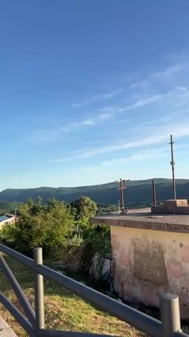The video captures a serene panoramic view of Pelinovo, Montenegro, from a slightly elevated vantage point. It opens with a static shot focusing on a small, weathered concrete building with a flat roof, topped with several metal poles or antennas. A dark metal fence runs across the bottom of the frame in the foreground.

The scene is set against a backdrop of lush green hills and mountains under a bright blue sky with scattered wispy white clouds, indicating clear and sunny weather. The lighting suggests late afternoon or early evening, with a warm, soft glow illuminating the landscape.

As the camera slowly pans to the right, it reveals more of the expansive mountainous terrain and a valley below. A small village or settlement becomes visible in the distance, nestled among the hills, with scattered houses and other buildings. A yellow van is briefly seen parked on a dirt patch near the concrete building, and a wheelbarrow is also visible. The video ends with a wider shot of the valley and the distant village.

No people are visible, and there is no discernible text within the frame. The video primarily focuses on the natural beauty of the mountainous region and a glimpse of local, rural architecture and infrastructure.