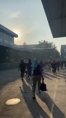 A group of people, predominantly women wearing headscarves, are walking away from the camera down a paved pathway. The sun is low in the sky, casting long shadows and creating a hazy, backlit effect. On the left is a multi-story building with several windows. On the right, a portion of another building is visible, with a dark, angled roofline. The pathway has painted lines and some reflective puddles. The people are dressed in casual attire, and some are carrying bags. The overall atmosphere is one of people moving from one location to another, possibly arriving at or departing from work or an event.