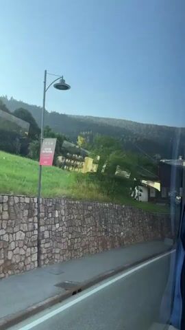 The video shows a view from a moving vehicle, likely a bus or car, looking out a window. The scenery is of a mountainous region with lush green hills and trees under a clear blue sky. A paved road runs parallel to the vehicle, bordered by a stone retaining wall and grass. On the other side of the road, houses with wooden accents and balconies are visible, nestled into the hillside. Some of the houses have signs, including one that appears to be for a hotel. The vehicle appears to be traveling through a small town or village. The reflections on the window distort some of the scenery, creating a shimmering effect. The time stamp indicates the video is a short clip from 00:00 to 00:04.