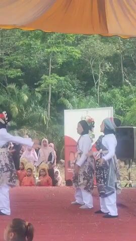 A group of people are on a stage performing a dance. Two young women in the foreground are wearing white tops and patterned skirts. They are both wearing headscarves and appear to be dancing in unison. In the background, several other people are watching the performance, including some children sitting on the stage. The stage is red and is set up outdoors with a backdrop of trees. There is a large orange tent over the stage.