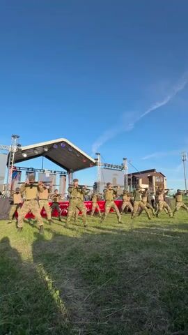 Against a bright blue sky, a group of about fifteen uniformed soldiers performs a choreographed dance with rifles on a grassy field. They are facing the camera and standing in front of a temporary stage structure with a small roof and visible lighting fixtures. Behind the stage, a large banner with text in a foreign language is visible. A modern-style house and a telecommunications tower can be seen in the background. The soldiers are wearing camouflage uniforms and caps, and they are holding their rifles in various positions as they move. The scene is bathed in sunlight, casting shadows on the grass.