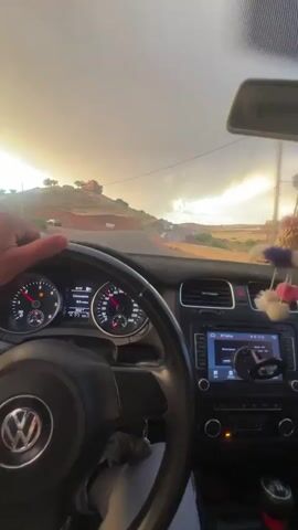 The video shows the interior of a car, specifically focusing on the driver's perspective. The driver's hands are visible on the steering wheel. The dashboard includes the car's speedometer, fuel gauge, and an infotainment system displaying a navigation interface. A decorative item is hanging from the rearview mirror. Outside the windshield, the view shows a paved road leading through a landscape with low hills and buildings. The sky is overcast, with sunlight breaking through the clouds. Another vehicle is visible on the road ahead. The camera's position is fixed, showing the driver operating the vehicle on the road.