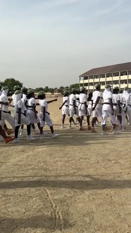 A group of people dressed in white shirts, shorts, and green hats are marching in formation on a dirt field. A man in a camouflage uniform stands to the side, observing them. There is a two-story building in the background. The weather is sunny. There is a dust cloud forming from the marching.