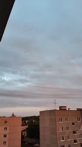 The video shows a view from a balcony looking out at an apartment complex. The sky is overcast with some pinkish hues, suggesting it might be sunset or sunrise. There are a few faint streaks that could be a rainbow. The buildings are multi-story residential apartments, some with balconies. Cars are parked in front of one of the buildings. Trees and other vegetation are visible in the background. The overall impression is a somewhat gloomy urban landscape.
