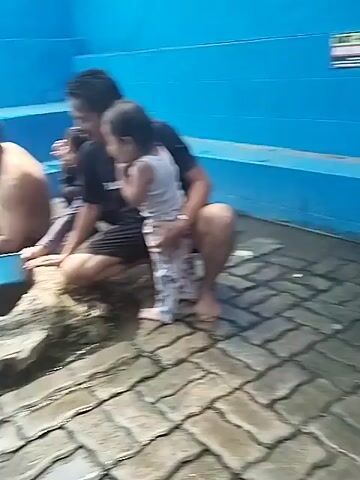The video shows a group of people gathered around a stone well. The well appears to be outdoors, possibly in a public area, with blue tiled walls in the background. Some people are sitting or crouching near the well, while others are standing. One man, shirtless and wet, is reaching into the well with a blue bucket. Another man is sitting beside him, also wet. A woman in a hijab is standing and holding a dipper. A child is standing near the woman. The ground is paved with interlocking stones, and there are some puddles of water. There are also signs visible on the blue wall. The overall impression is of a communal gathering at a water source.