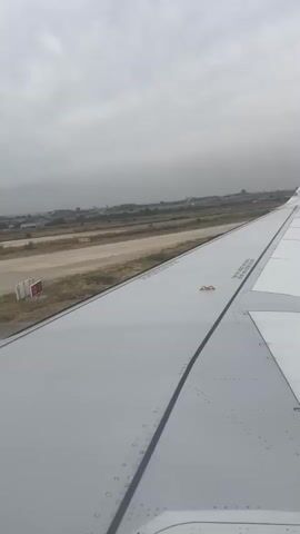 The video shows the wing of an airplane from the perspective of a passenger. The airplane is on the ground at an airport, with a runway visible in the background. The sky is overcast. The wing is white with a blue stripe at the tip. There are markings and rivets on the wing. The runway is paved and there are some buildings and vegetation in the distance. The airplane appears to be stationary.