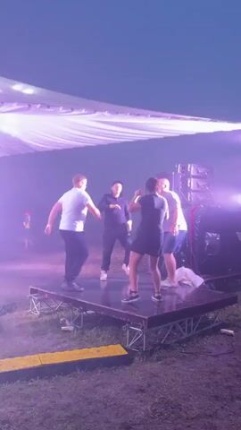A group of four people are dancing on a small stage outdoors. The stage is made of metal scaffolding and a dark surface, and is covered by a large white canopy with yellow lights. To the right of the stage, there are large black speakers stacked on top of each other. The ground around the stage is covered in grass and debris. In the background, there is a hazy, overcast sky and another person can be seen walking in the distance. The people on the stage are dressed casually and appear to be enjoying themselves. The lighting changes from white to purple and then back to yellow throughout the video, creating a dynamic atmosphere.