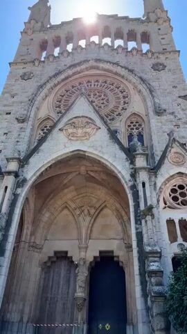 The video opens with a low-angle shot of a large, old, stone church with ornate architectural details, including a large rose window. The camera pans up to reveal a clear blue sky. The camera then moves to show a multi-story building with a peach-colored facade, adorned with flags and banners. The camera continues to pan, showing a town square with trees, buildings, and people walking around. The sky remains clear and blue. A woman wearing sunglasses is seen briefly in the foreground.