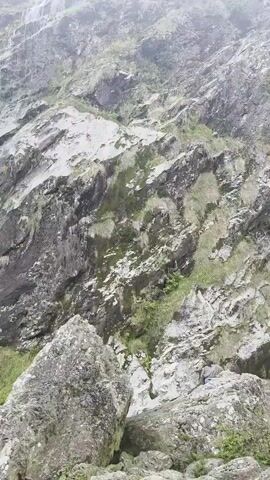 The video shows a person wearing a yellow vest standing on a rocky outcrop in front of a cascading waterfall. The waterfall flows down a steep, rocky mountainside covered in moss and vegetation. The surrounding environment is misty and appears to be a natural landscape. The person is facing the waterfall, possibly admiring the view.