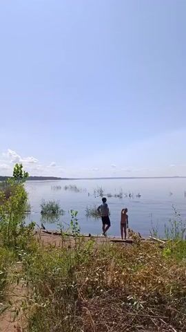 A bright sunny day unfolds over a calm lake. The sky is a clear, pale blue, with a few wisps of white clouds on the horizon. The water is a serene, reflective surface, disturbed only by small ripples. Tall grasses and reeds grow along the shore, some dry and brown, others vibrantly green. In the foreground, a path of dirt and dry vegetation leads towards the water.

Three people are visible near the shore. On the left, a person wearing a dark shirt and light shorts stands facing the water. Next to them, a child in a yellow swimsuit stands with their back to the camera. Further out in the water, a third person, silhouetted against the bright sky, wades with a fishing net or similar equipment. The person with the net appears to be fishing or gathering something from the water. The scene conveys a sense of peaceful outdoor activity.