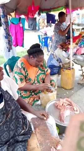 A busy market scene. Three women are visible. One woman is in the background, wearing a black and white patterned dress. She is seated and looking down. In the middle of the frame, a woman wearing an orange and green patterned dress is seated and holding a small, white object in her hands. She is looking down at the object. In the foreground, a person wearing a black and white patterned top is seated and looking towards the woman in the middle. They are near a large cutting board with a meat cleaver. Behind the seated women, there are various items for sale, including clothing and yellow containers. The ground appears to be dirt or gravel. The scene is lit by natural light.