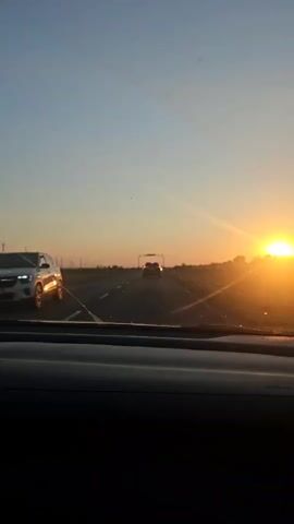 The video captures a first-person perspective from inside a car, looking out the windshield. The scene is a highway at sunset, with the sun positioned low on the horizon, casting a warm, golden glow across the sky and the road. The sky transitions from a light blue at the top to a pale orange near the horizon.

The primary subjects are the road and the sunset. In the distance, a single car is visible driving ahead, its taillights illuminated by the fading light. There is also an overhead gantry or sign structure on the highway. The foreground shows the dashboard of the car, with a dark, textured surface.

The activity is simply driving on a highway. The time of day is clearly sunset, indicated by the sun's position and the warm, diffused lighting. The weather appears clear and calm, with no visible clouds. While the exact location is identified as Көксаяқ ауылдық округі, Kazakhstan, this is inferred from external information and not directly visible in the footage. The overall mood conveyed is one of a peaceful journey or a scenic drive during dusk. There is no visible text in the video.
