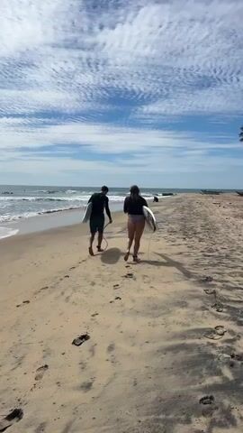 Two people, one male and one female, walk away from the camera along a sandy beach, each carrying a surfboard. They are walking towards the ocean, which has moderate waves and appears to be populated with other surfers. The sky is blue with scattered white clouds. The scene is a bright, sunny day at the beach.