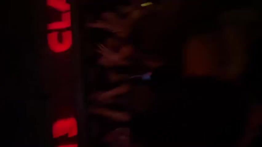 The video is a dark, vertical shot of a group of young adults appearing to be at a club or party. A bright red neon sign is visible vertically on the left side of the frame, displaying the text "CLASS 313" in large block letters. Some individuals in the foreground and mid-ground are looking towards the camera and smiling, with one person making a gesture with their hand. The lighting is dim, with the neon sign being the primary light source, creating a somewhat blurry and atmospheric scene. It is difficult to discern specific actions or interactions beyond general socializing and awareness of the camera.