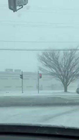 The video shows a snowy scene viewed from inside a car. A street covered in snow is visible, with a large, bare tree on the right. In the background, across the street, there is a light-colored, one-story building with a flat roof. Two traffic lights are visible; one has a red light illuminated. The sky is overcast, and snow is falling. A "RIGHT ONLY" street sign is seen above the street.