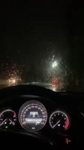 The video shows the interior of a car at night during a heavy rain. The car is stationary, as indicated by the speedometer reading 0 km/h. The dashboard is visible, displaying various gauges and indicators. Outside the windshield, streetlights and other car lights are blurred by the rain.