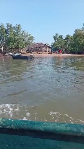 The video depicts a boat moving across water on a sunny day. On the shoreline to the left and right are trees and what appear to be houses. On the shore to the right, there are at least two people visible, one wearing a red shirt. The boat is moving and leaving a white wake in the water behind it.