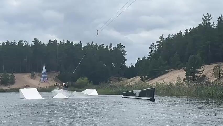 A person is wakeboarding on a lake, being pulled by a cable system. They are wearing a helmet and shorts. In the background, there is a wooded area with sand dunes and a cloudy sky. There are also several obstacles in the water, including ramps and a rail. The wakeboarder is kicking up a large spray of water as they ride. The video pans across the scene, showing the wakeboarder from different angles.