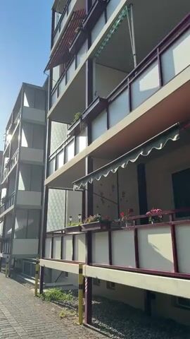 The video shows a view of an apartment building with balconies and a paved walkway in the foreground. The sun is shining brightly, casting shadows on the building. There are cars parked along the walkway, and some trees and other buildings are visible in the background. The overall scene is one of an urban environment on a sunny day.