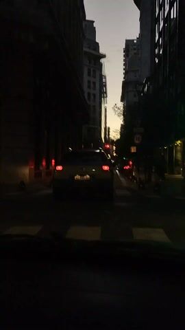 The video shows a street scene at dusk from the perspective of a car. The car is stopped at a crosswalk behind another car. Tall buildings line both sides of the street, casting long shadows. The sky is visible between the buildings, showing the soft colors of the setting sun. Red taillights of cars ahead can be seen, and a street sign with the number 10 is visible on the right. The overall scene is dark, with limited lighting.