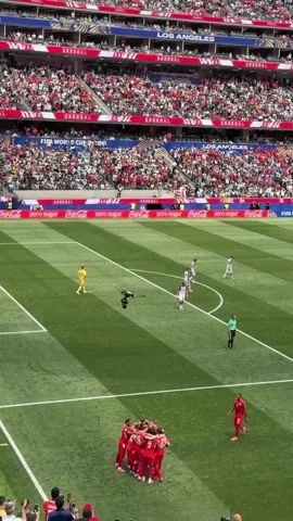 The video shows a soccer match from a high angle, looking down onto the field and into the stadium crowd. The field is green with white lines marking the boundaries and penalty areas. In the foreground, a group of players in red uniforms are huddled together, celebrating. There are other players in white uniforms on the field, as well as a referee. In the background, a large stadium filled with spectators is visible, with red and white seating. Advertising banners are displayed around the stadium, including Coca-Cola. A large screen at the top of the stadium shows "LOS ANGELES". The overall impression is of a lively and energetic sporting event.
