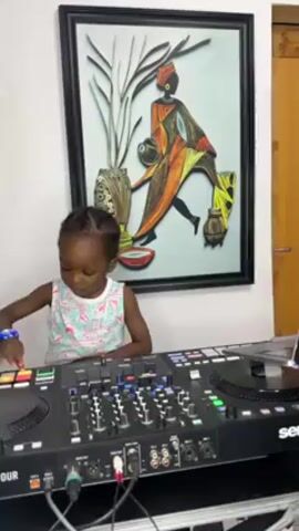 A young child stands in front of a DJ controller and presses buttons. The child has dark skin, and is wearing a floral tank top and a blue beaded bracelet. Behind the child is a painting with a black frame, that features an abstract image of a woman, carrying things on her head and back. The DJ controller is black, with many buttons and sliders. Wires are visible under the controller. A doorway is visible to the right of the painting.
