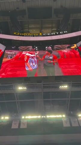The video shows a large screen in an arena displaying two people talking. The screen is suspended from the ceiling of the arena, which has a Golden 1 Center sign visible at the top. Below the screen, a monster truck is parked. The monster truck is red and white with large tires. The screen shows a man in a red and white shirt and a woman in a red and black shirt. On either side of the main screen, smaller screens show the face of the man. The lighting in the arena is dim, with spotlights illuminating the monster truck and parts of the arena. The overall impression is of a monster truck event in an indoor arena.