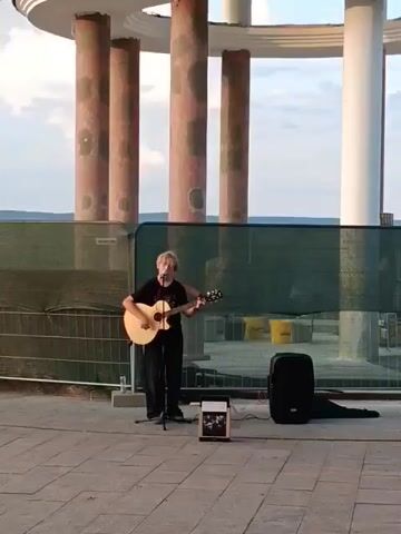 A person stands in front of a building with large columns and a circular roof, playing an acoustic guitar. The person is wearing a dark shirt and pants and has short, light-colored hair. They are also wearing a microphone and have a small amplifier and a speaker next to them. The background shows a fence and some construction materials. The sky is visible in the background.
