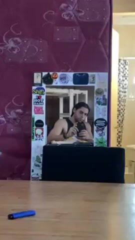 A man takes a photo of himself in a mirror. The man has long dark hair pulled back from his face and wears a black tank top. The mirror is surrounded by stickers. A table is in the foreground, with a blue object on it. Behind the mirror is a red mattress. The background shows a room with a bathroom visible in one corner.
