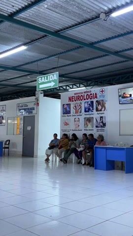 The video shows a waiting area in a medical clinic. Seven people are sitting in chairs in front of a blue reception desk. Behind them is a poster that says "Servicio Medico Neurologia". A television is mounted on the wall above the poster and displays a video of a doctor talking. Above and to the left, a green sign indicates "Salida" (Exit). The area is brightly lit by overhead fluorescent lights. The floor is covered with white square tiles. A sign for "Traumatologia y Ortopedia" is visible to the left of the "Salida" sign. The room appears clean and orderly.