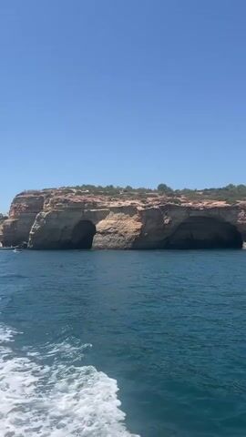 The video captures a wide shot of a bright, sunny day with a clear blue sky. The foreground shows the wake of a boat on the deep blue ocean. In the middle ground, several kayaks with people paddling are visible. The background features a dramatic coastal cliff face with natural arches and caves carved into it. Vegetation covers the top of the cliffs.