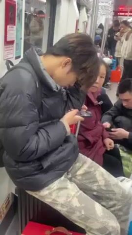 The video shows a young man in a black puffer jacket and camouflage pants sitting on a red suitcase on a crowded subway car. He is absorbed in his phone. There are other passengers nearby, some seated, and some also looking at their phones. A red fire extinguisher is visible on the wall of the subway car.  The car is moving and appears to be quite full.