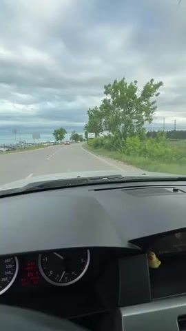 The video shows a car's-eye view of a drive on a road on a cloudy day. The car is traveling on a road alongside a body of water, with a few cars parked along the shoreline. There is green vegetation along the side of the road. The driver is visible from the side, and the car's dashboard is visible. The view of the road ahead is seen through the windshield, and the rearview mirror shows the road behind. The side mirror shows another car following behind. The driver seems to be accelerating as the car speeds up. The steering wheel is black, as are the interior panels of the car. A small, yellow rubber duck sits on the dashboard near the infotainment screen. The speedometer and other gauges are visible in the instrument cluster.
