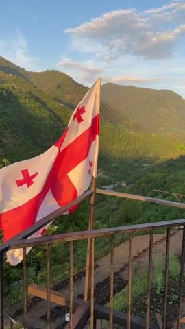 The video shows the flag of Georgia, a white flag with a large red cross in the center and four smaller red crosses in the four quadrants, fluttering in the wind. The flag is attached to a pole on a balcony with a metal railing. Behind the flag is a view of green, forested mountains under a blue sky with some wispy clouds. The light suggests it is late afternoon or early evening. The balcony has a small garden bed. The camera is steady.