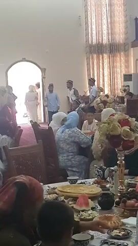 The video shows an indoor gathering, possibly a celebration or wedding, with people seated around tables laden with food. In the background, a doorway leads to another area where several individuals are standing. One man in the foreground, wearing a light blue shirt and a patterned skullcap, is holding a long, brass musical instrument, possibly a horn or trumpet, and appears to be playing it or preparing to play it. He is looking towards the doorway. Behind him, two other men in similar skullcaps are also standing; one is holding a drum. Several women and children are seated at the tables, some looking towards the musicians. The overall environment suggests a festive occasion.