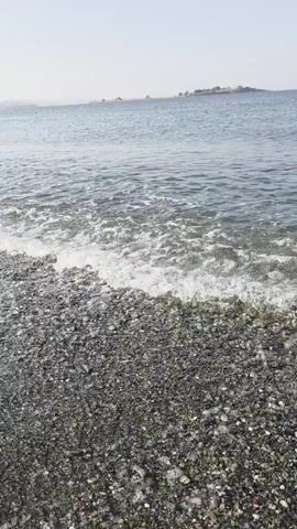 The video captures a close-up, low-angle view of a pebble beach with gentle waves lapping at the shore. The water is clear, showing small pebbles beneath the surface as the waves recede. The beach is composed of dark, smooth pebbles. In the background, the sea stretches towards the horizon under a hazy sky. Distant landmasses or structures are faintly visible on the horizon. The video focuses on the interaction between the water and the shore, emphasizing the texture of the pebbles and the movement of the waves. There are no people or other discernible objects in the immediate foreground or background. The overall impression is one of a calm, natural coastal environment.