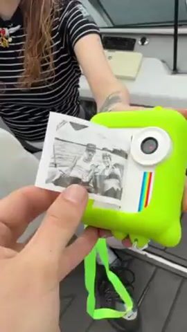 A young woman with long curly hair, wearing a black and white striped shirt, white tights, and black shoes, stands on a boat holding a green toy camera with a photo sticking out of the top. A person, whose face is not visible, is holding the photo and camera, and looking at the photo. The photo is a black and white image of two people in a boat. The camera has a white square on the front with a rainbow stripe and a lens above it. The boat has a white interior, and a gray floor. The sound of wind is audible.