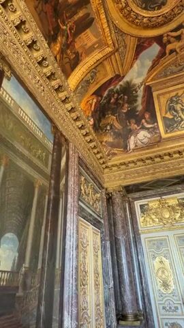 The video shows a panning shot of an opulent interior, likely a palace or grand hall. The ceiling is heavily decorated with intricate gold detailing and large, detailed frescoes depicting mythological or historical scenes, including cherubs, figures in togas, and elaborate landscapes. Large marble columns with gold accents support the ceiling. The walls also feature large paintings and ornate gold trim. In the lower portion of the video, ornate doors with gold detailing are visible, as well as a statue in an alcove. The lighting is warm and appears to be artificial, creating a sense of grandeur and richness. No people are clearly visible or interacting in the scene.