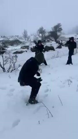 Three people are having a snowball fight in a snowy, foggy environment. They are all wearing dark winter clothing. The person in the foreground is crouched down, preparing to throw a snowball. The person in the middle is standing with their arms outstretched. The person on the right is also standing, seemingly ready to throw. There is a chain-link fence behind them, with some vegetation peeking through the snow. The overall scene appears to be a winter landscape with heavy snowfall.
