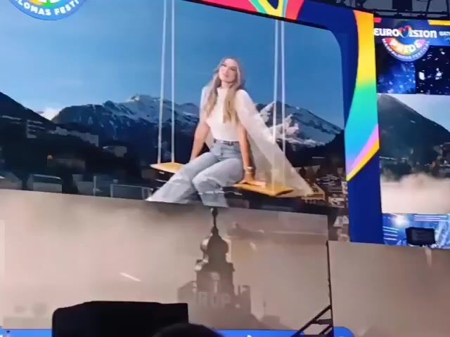 The video shows a large screen displaying a woman on a swing in front of a mountain village backdrop, with the logo "EUROPRIDE MASPALOMAS FESTIVAL" at the top. The screen then transitions to a graphic that reads "GREETINGS FROM AUSTRIA TO Australia", also featuring a mountain village scene. The bottom of the screen is lined with spotlights. There appears to be smoke or haze near the base of the stage. The video seems to be taken from the audience perspective, focusing on the stage display.