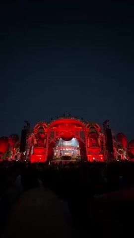 A vertical, wide shot captures a dimly lit outdoor music festival at night. In the foreground, the silhouettes of a crowd of people are visible, facing a large stage. The stage is illuminated with vibrant red lights and features an elaborate, ornate structure with a domed roof. Red lights also emanate from large, circular structures positioned on either side of the stage, resembling inflated balloons. The sky is dark and clear. The overall atmosphere is one of excitement and festivity.