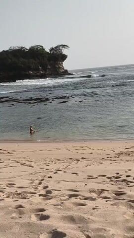 A person is standing in the shallow water at the edge of a sandy beach. The ocean waves are breaking in the background, and a rocky island with trees is visible offshore. The sky is clear and overcast.