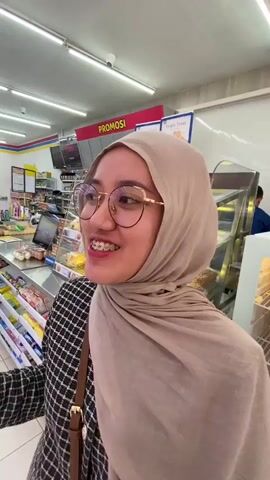 The video shows a young woman wearing a beige hijab and glasses, with braces on her teeth. She is in a convenience store, possibly a minimarket, with shelves of products visible in the background. She is smiling and sticking her tongue out at the camera. The lighting suggests an indoor setting.