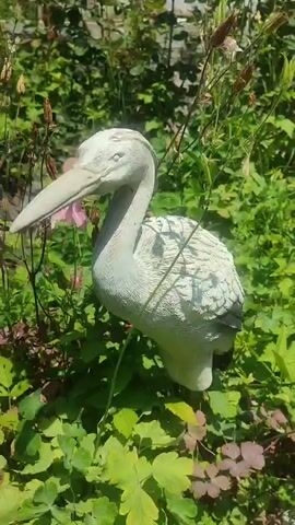 A bird statue stands in a garden among green plants. The statue is white and textured, with a long beak and speckled body. The surrounding plants are lush and green, with some pink flowers visible in the background. The sunlight casts shadows on the scene.