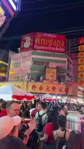 The video shows a crowded street at night, presumably during a festival or celebration in a Chinese community. There are many people walking around, dressed in various outfits, some in traditional Chinese attire. Red lanterns hang strung across the street, adding to the festive atmosphere. Buildings along the street are decorated with large signs written in Chinese characters and Thai script. There are signs in Thai script that appear to read "Tak Seng Rung". A large air cooler is visible in the foreground, indicating warm weather. Several people wearing sashes and crowns walk through the crowd. The overall atmosphere is lively and vibrant.