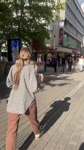 The video captures a bustling pedestrian street scene during daylight. In the foreground, a woman with blonde hair pulled back in a ponytail, wearing a gray and black plaid shirt and brown pants, walks away from the camera. She appears to be wearing headphones. Her shadow is cast on the cobblestone pavement. To her left, a large tree provides shade. The street is lined with multi-story buildings housing shops, including one with a prominent "SATURN" sign. Numerous people are walking in both directions, some carrying shopping bags. The sky is partly cloudy. The camera pans slightly to the right, revealing more of the street and the shoppers.