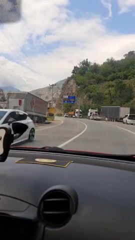 The video begins with a view from inside a vehicle, looking out at a road with several trucks and cars. A large white truck with red and blue lettering is visible on the left, with part of its trailer cut off by the frame. A yellow smiley face sticker is superimposed on the video. The camera then pans to the right, revealing a curved road with more trucks parked on the side. A blue directional sign is prominently displayed, with Turkish text indicating directions to "Şehir Merkezi" (City Center), "Kafkasör Yaylası" (Kafkasör Plateau), "Borçka", "Rize", "Trabzon", and "Batum". The road is flanked by a steep, rocky hillside on the right and buildings in the distance on the left. The sky is cloudy. As the video progresses, the camera continues to move forward, showing more of the traffic and the surrounding landscape. The buildings in the background appear to be a mix of residential and commercial structures, possibly a small town or city. The overall scene suggests a mountainous region with a busy highway.